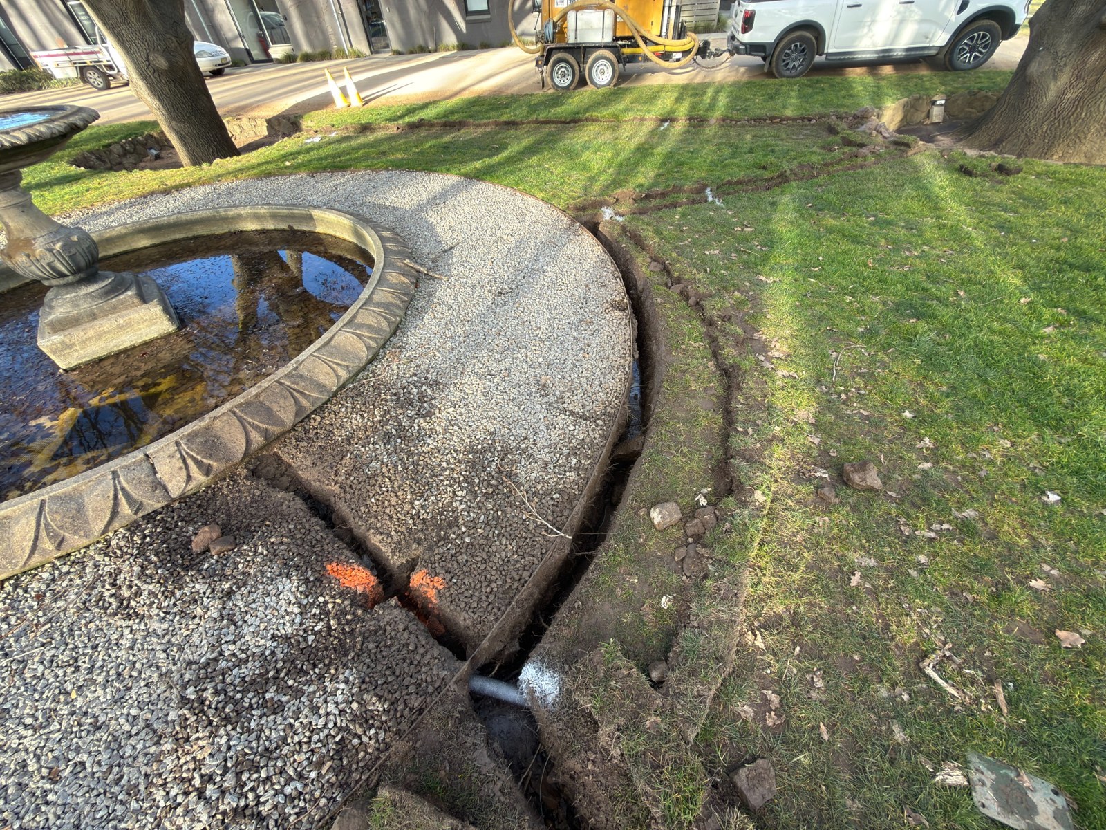 GreenVac trenching around a fountain and established trees, with the compact trailer parked nearby